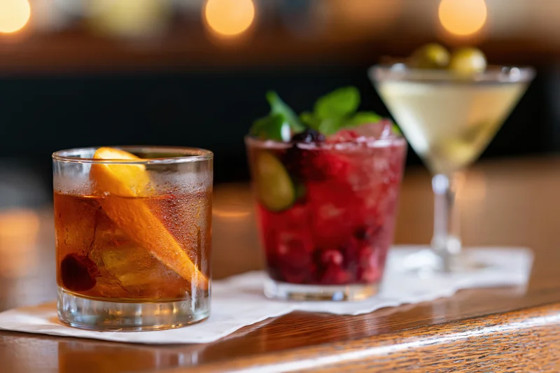 Three cocktails with garnishes arranged on a wooden bar top under warm lighting