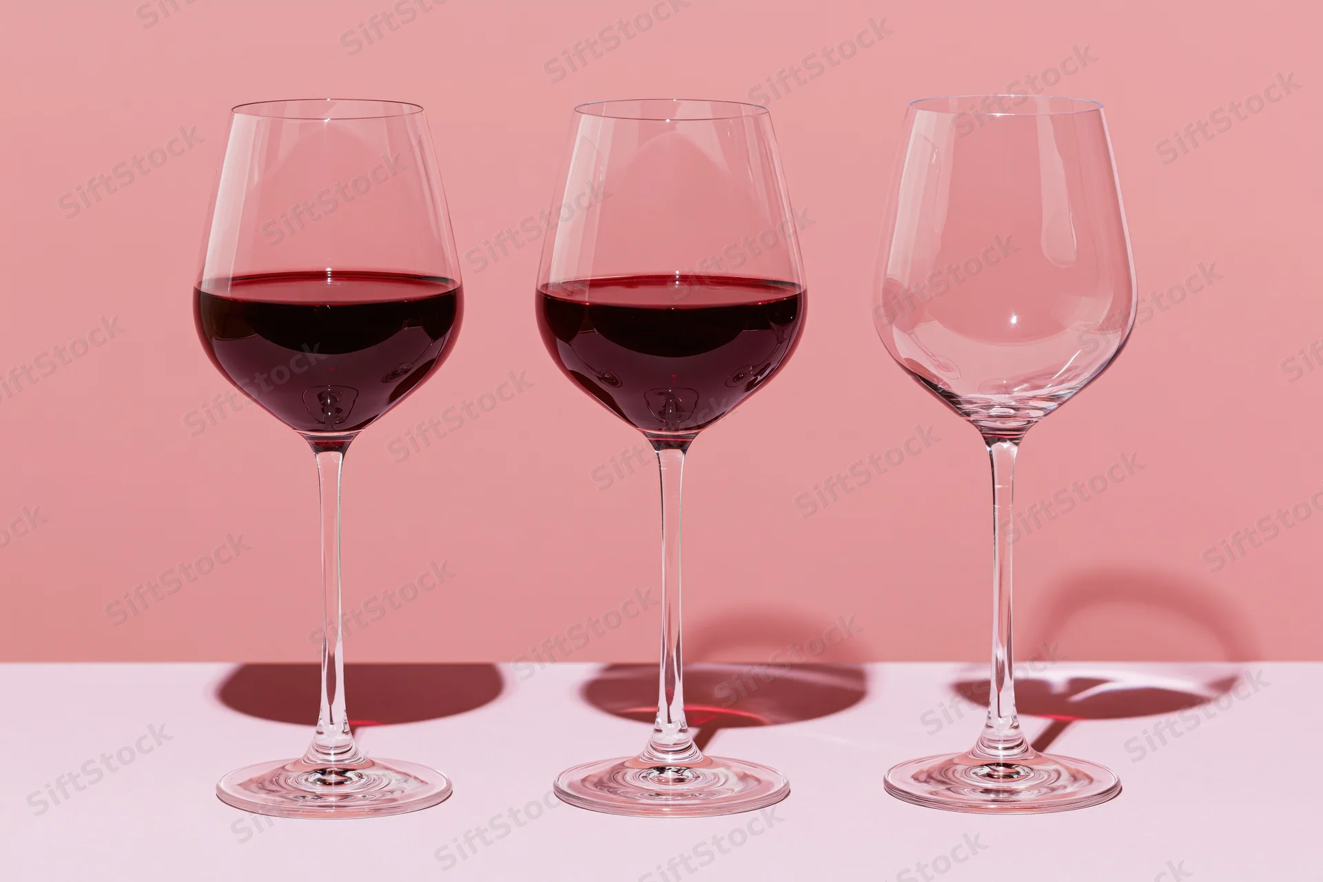 Three wine glasses in a row, two filled with red wine, one empty, on a pink surface