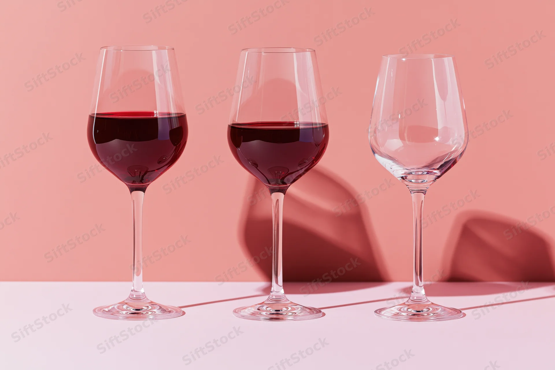 Three stemmed wine glasses in a row on a pink surface with shadows and reflections