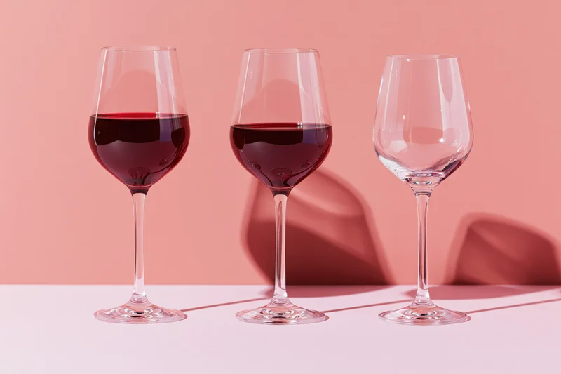 Three stemmed wine glasses in a row on a pink surface with shadows and reflections
