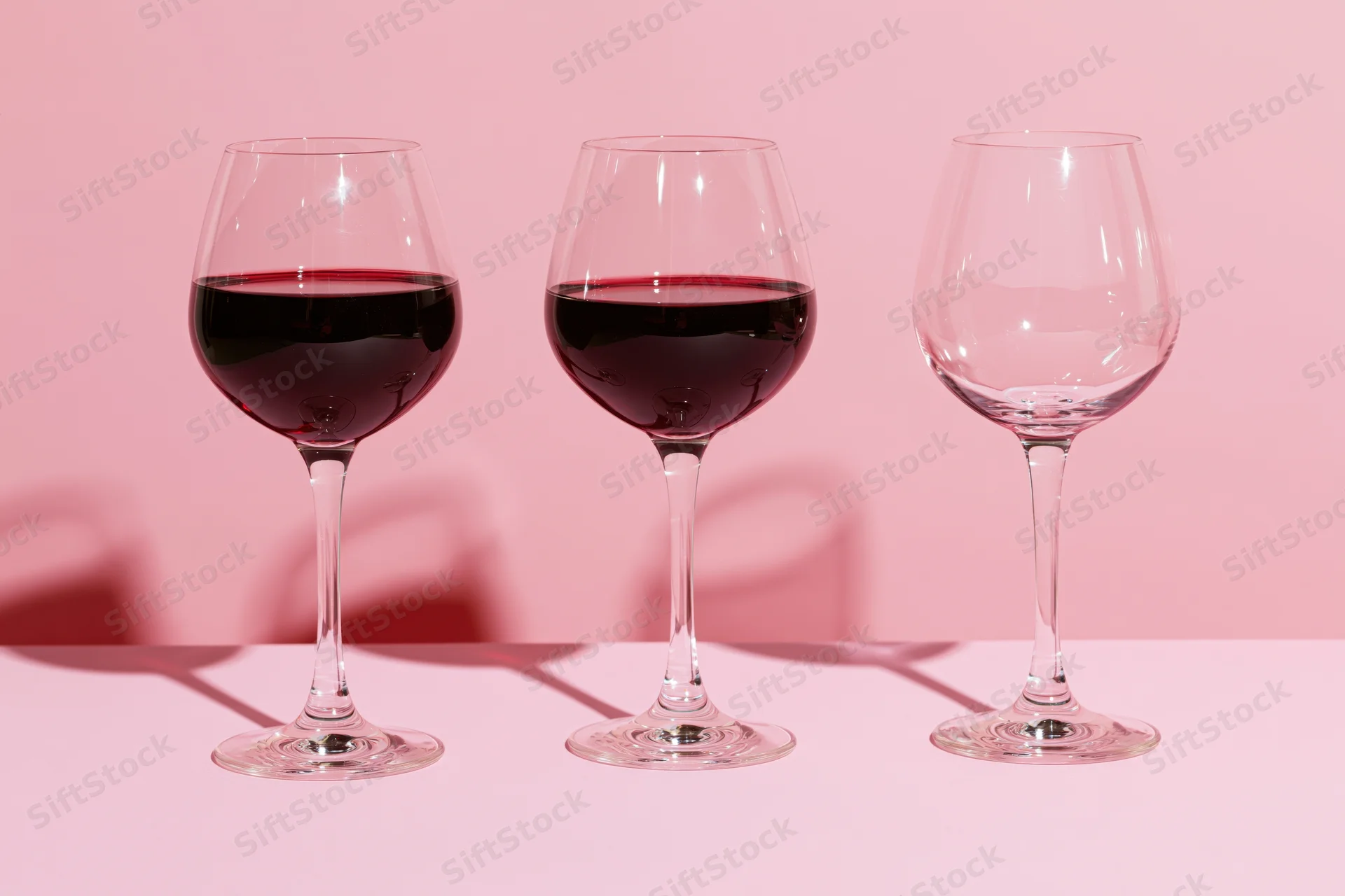 Three wine glasses in a row, two filled with red wine, one empty, on a pink surface