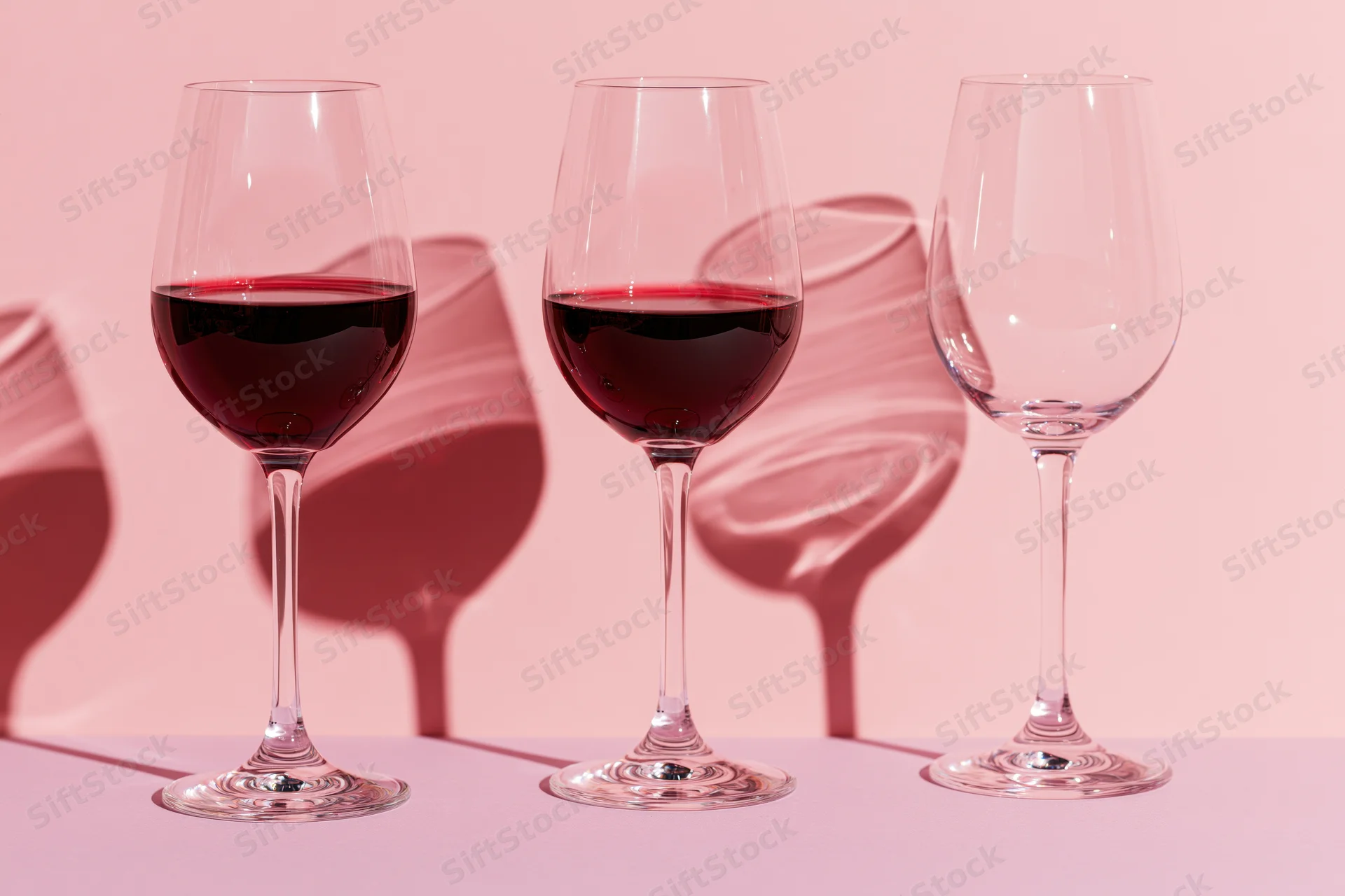 Three stemmed wine glasses in a row, two filled with red wine, on a flat pink surface with soft shadows