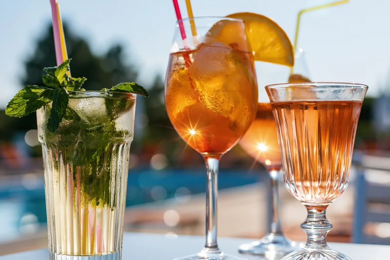 Three summer cocktails with mint, orange, and straws on an outdoor table by a pool