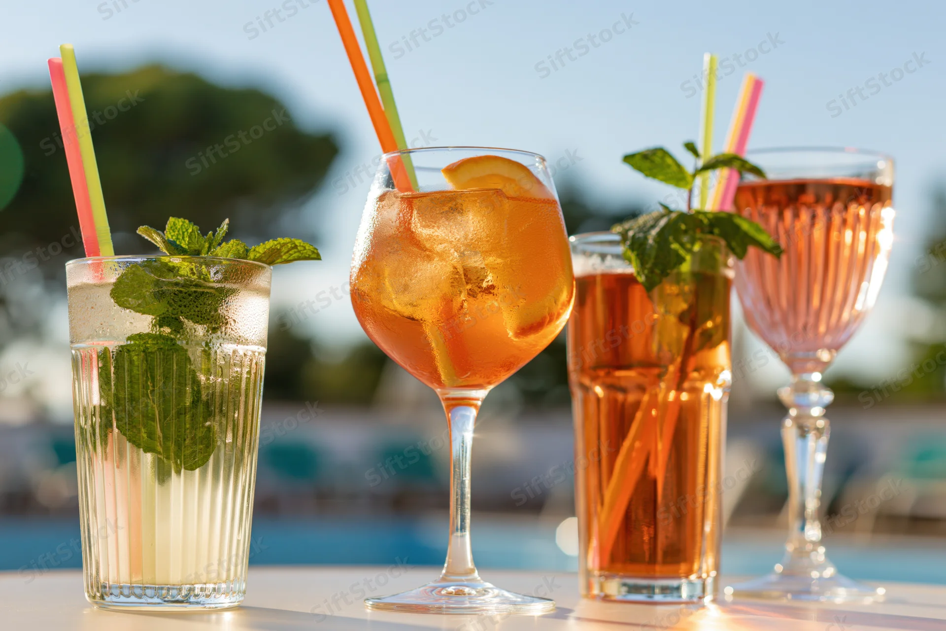 Four summer cocktails with mint and citrus garnishes on an outdoor table by a pool