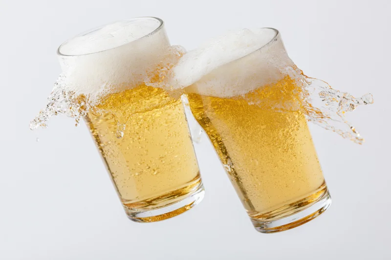 Two pint glasses of pale lager clinking with foam splashing against white background