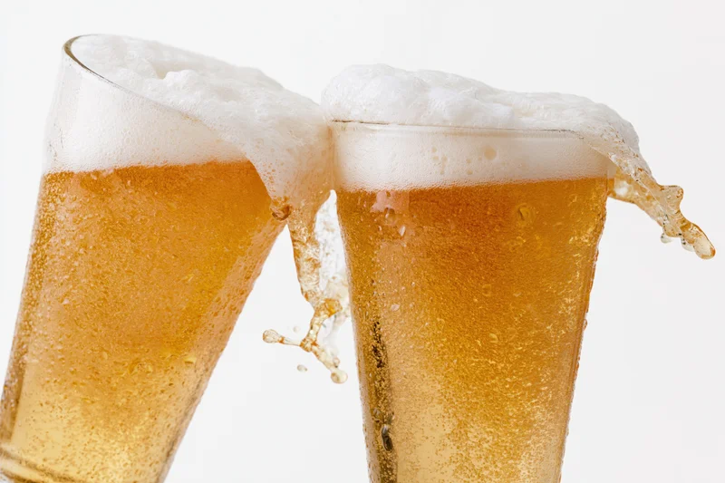 Two pint glasses of pale lager clinking together with foam spilling over the edges against a white background