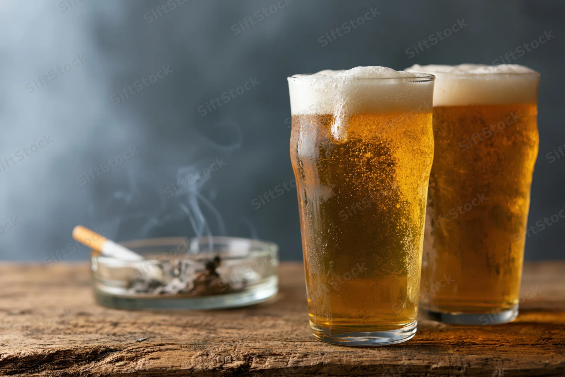 Two cold pint glasses of golden lager with foam heads on a wooden table beside a smoking cigarette in an ashtray