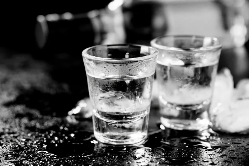 Two full shot glasses of clear spirit on a wet dark bar surface with condensation and ice nearby