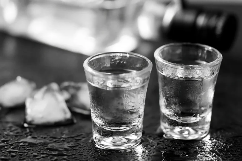 Two full shot glasses of clear spirit on a wet dark surface with ice and condensation