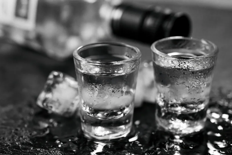 Two full shot glasses of clear spirit on a wet dark bar surface with ice and a bottle in the background