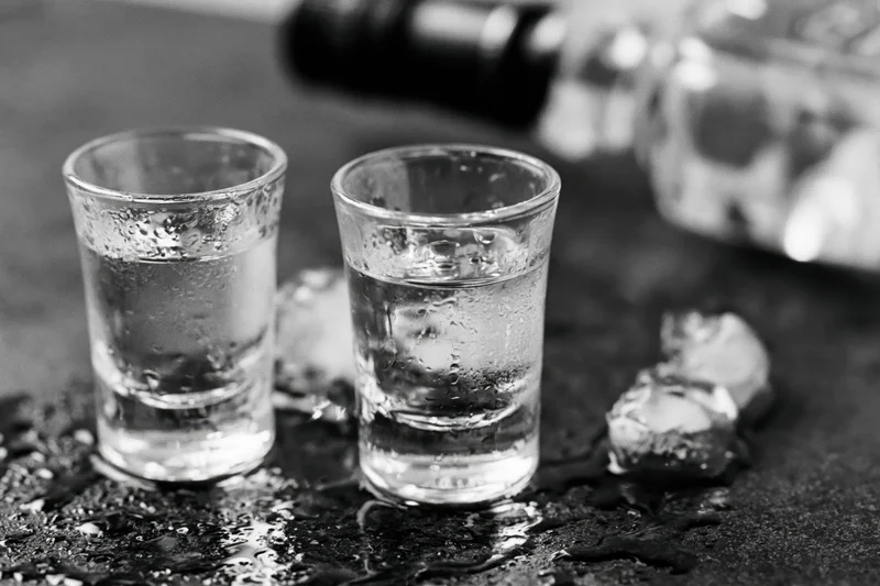 Two full shot glasses of clear spirit on a wet dark bar counter with ice cubes and condensation