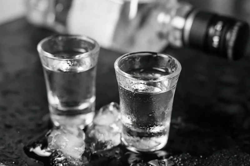 Two full shot glasses of clear spirit on a wet dark surface with ice and condensation, close-up view