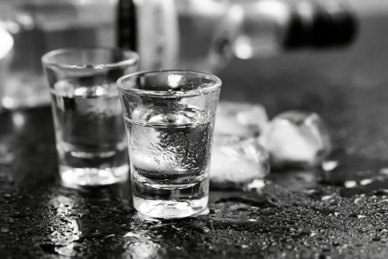 Two shot glasses filled with clear spirit on a wet dark bar surface with ice and condensation