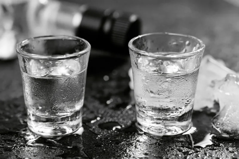 Two full shot glasses of clear spirit on a wet dark surface with condensation and ice nearby