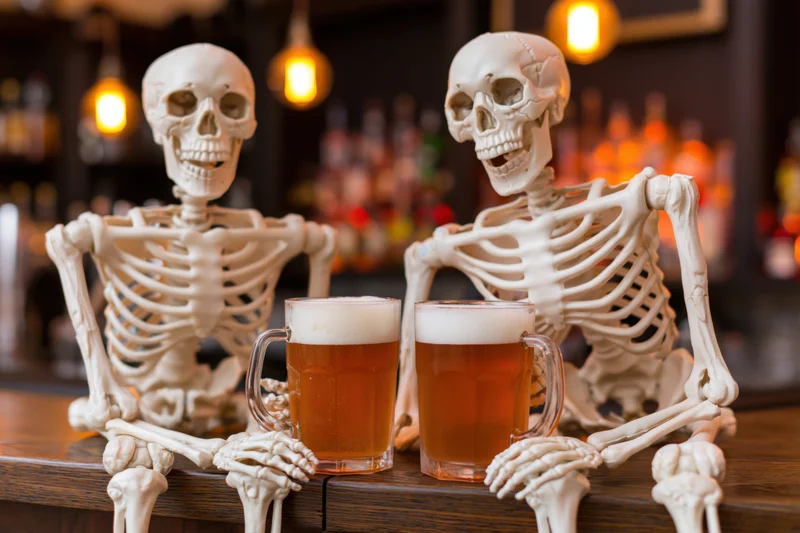 Two plastic skeletons sitting at a wooden bar with beer mugs in front of them