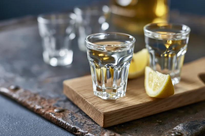Two shot glasses filled with clear liquor on a wooden board with lemon slices, set on a dark metal surface