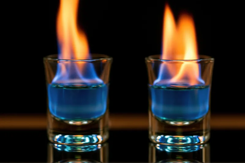 Two shot glasses with blue liquid burning in flames on a dark reflective surface