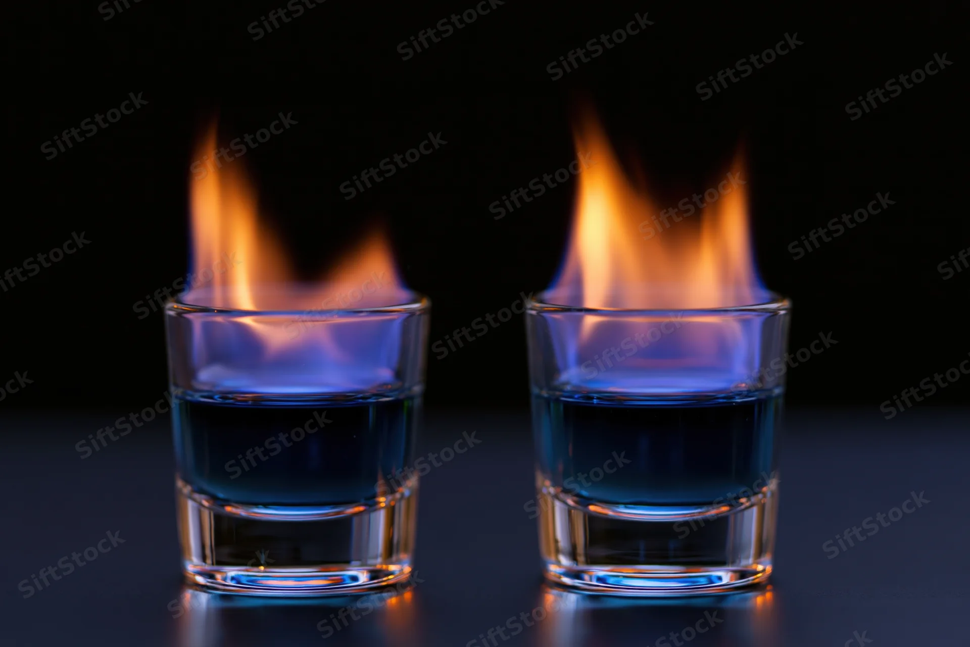 Two shot glasses with burning blue liquid flames on a dark reflective surface
