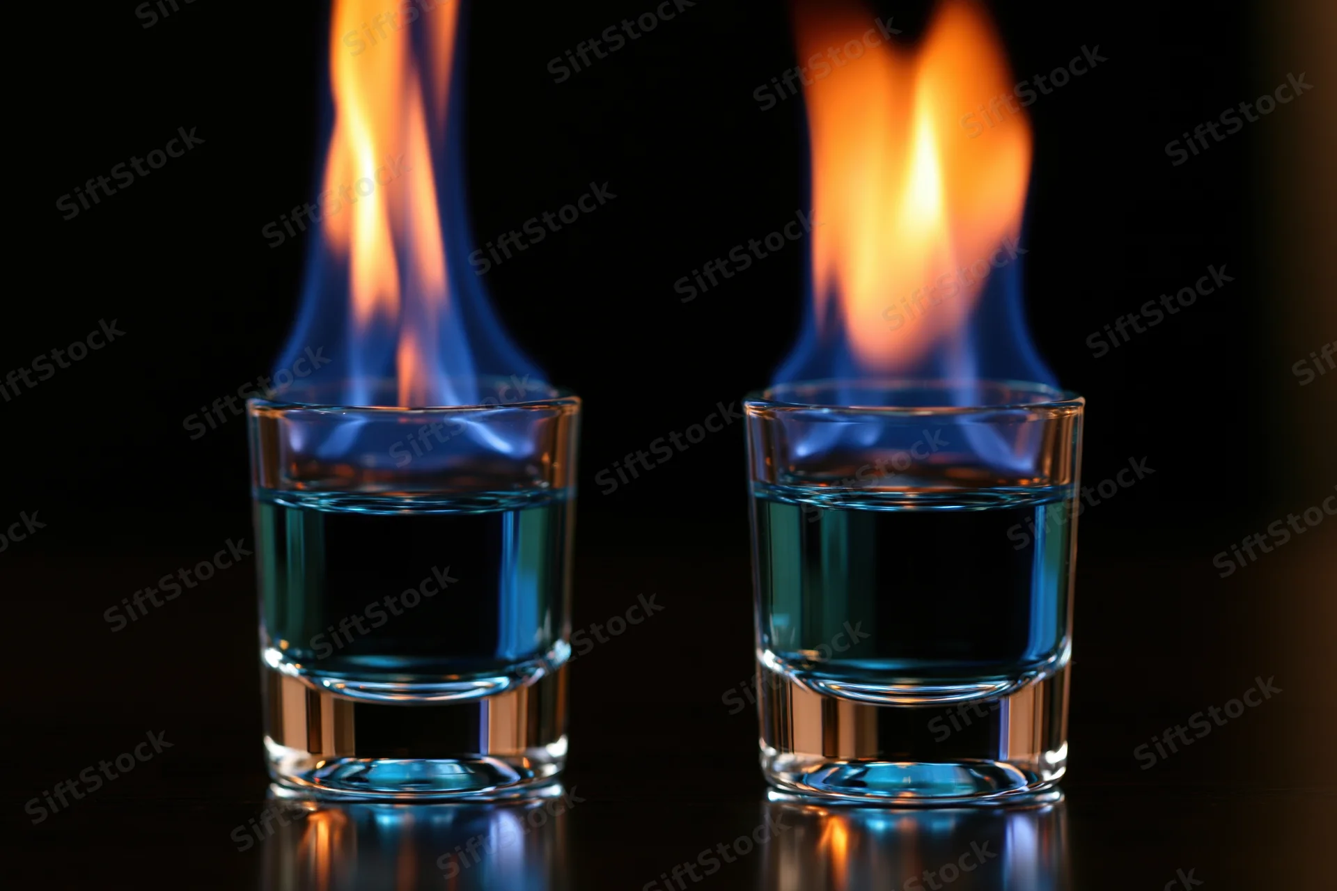 Two shot glasses with burning blue liquid flames on a dark reflective surface