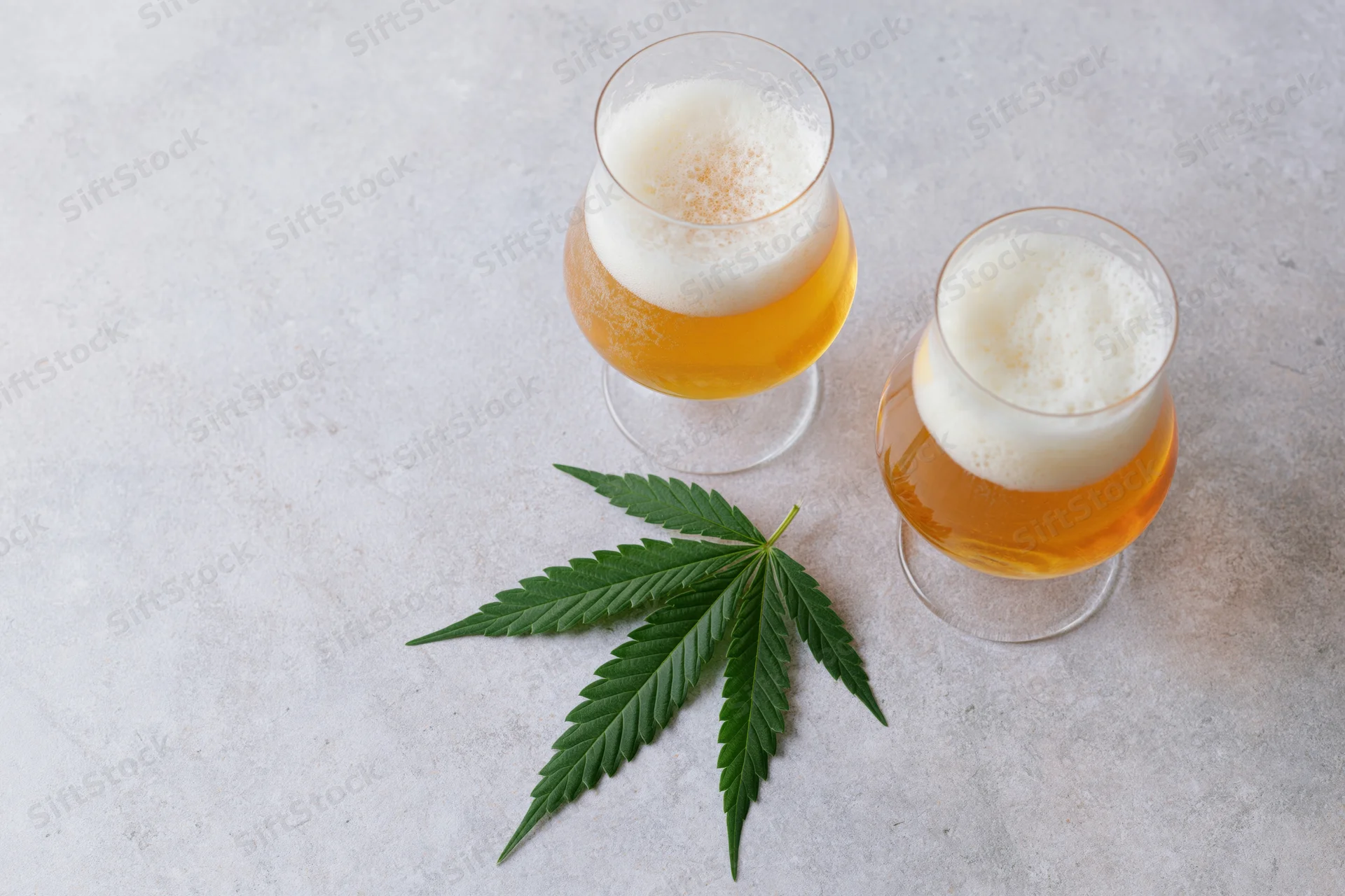 Two tulip glasses filled with pale golden beer topped with white foam beside a cannabis leaf on a light surface