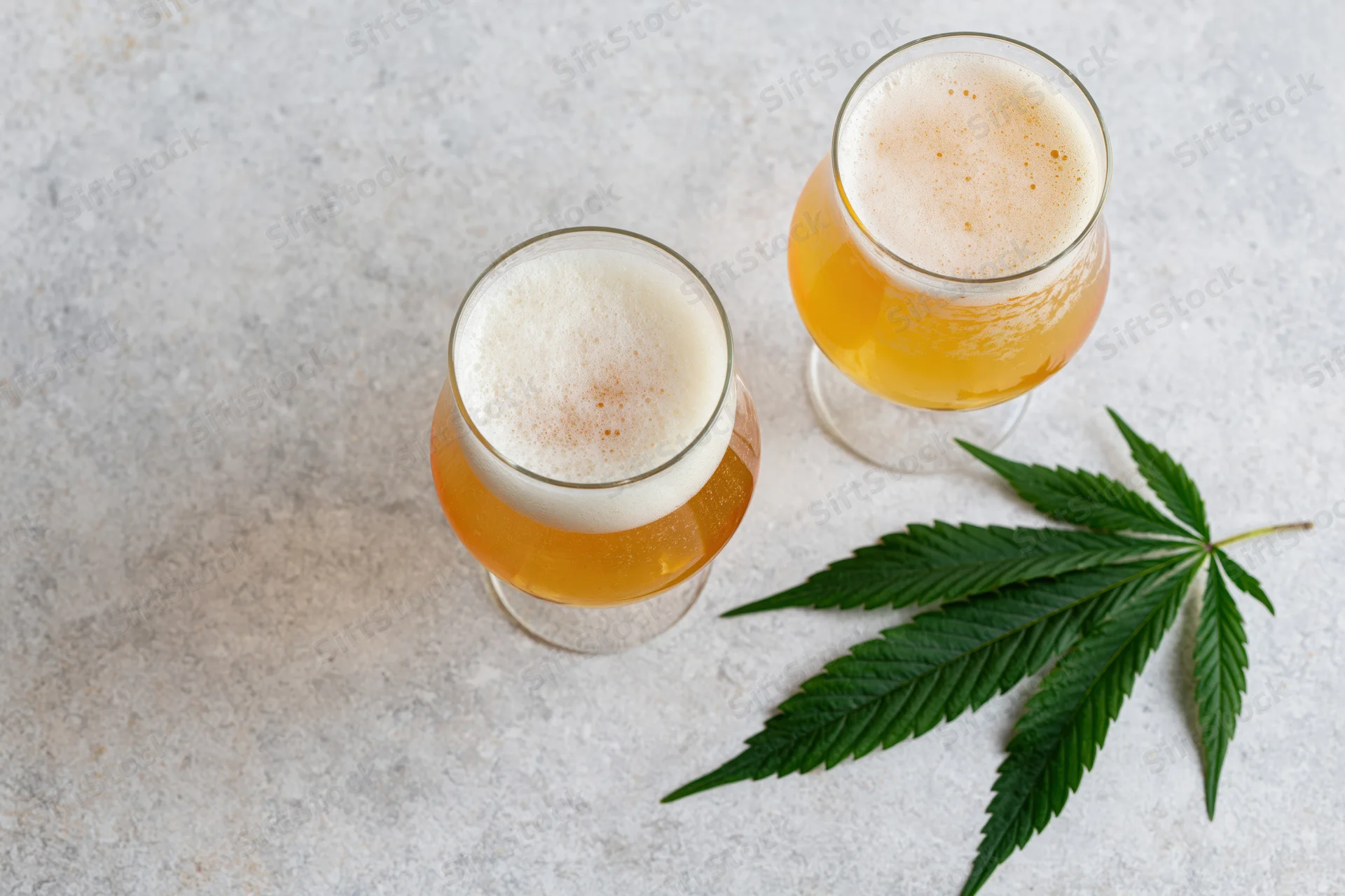 Two tulip glasses of pale golden beer with foam and a cannabis leaf on a gray concrete surface