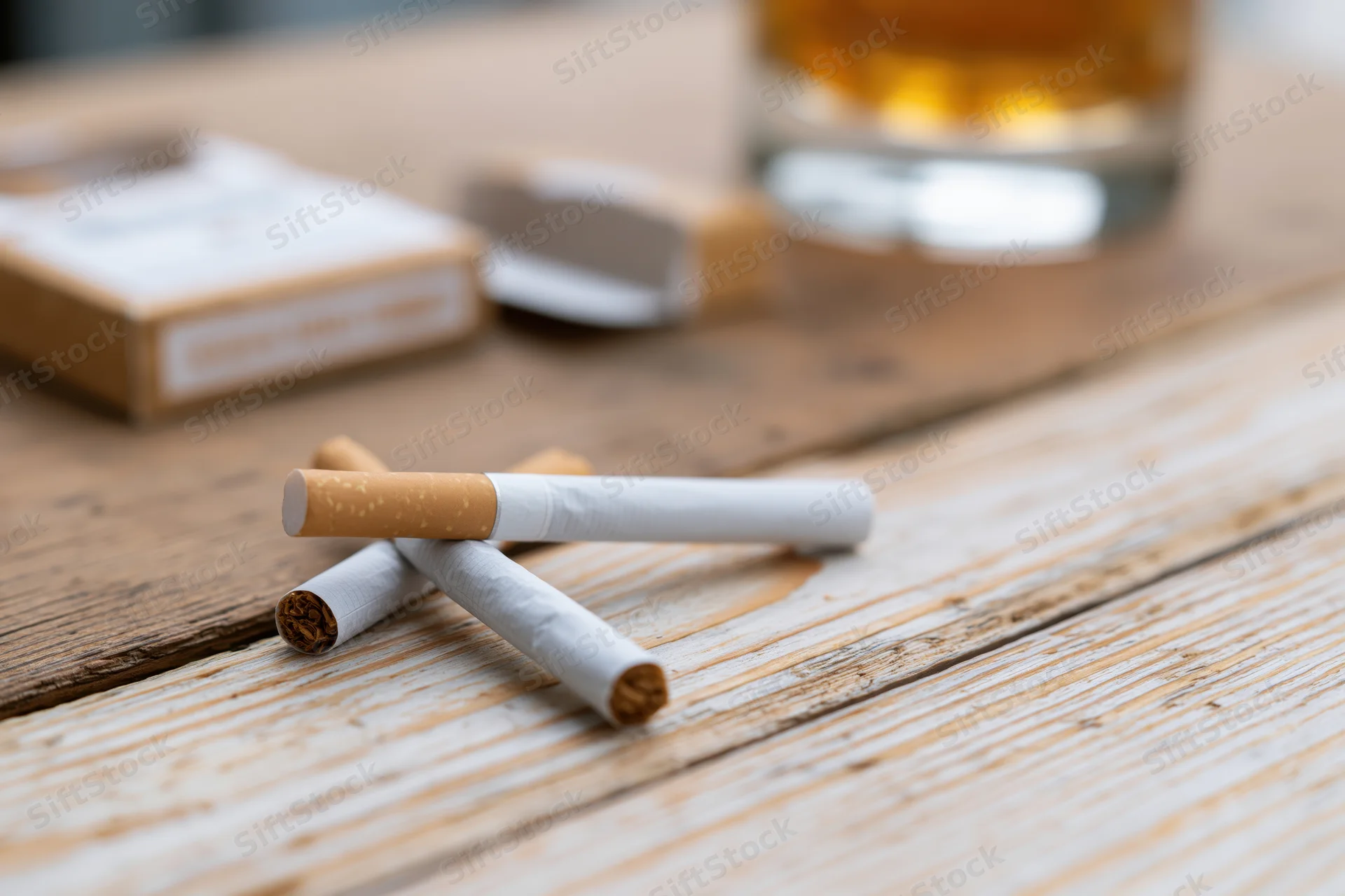 Two unlit cigarettes crossed on a worn wooden table with a glass of whiskey and an open pack nearby