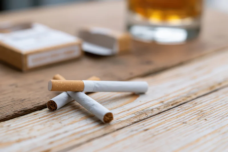 Two unlit cigarettes crossed on a worn wooden table with a glass of whiskey and an open pack nearby