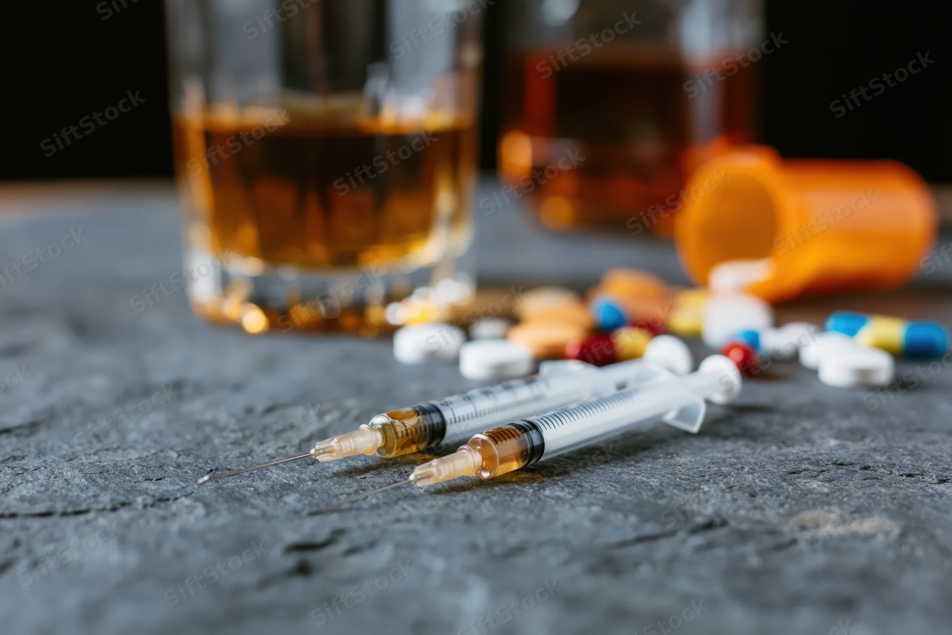 Two used syringes with pills and a glass of whiskey on a dark stone surface
