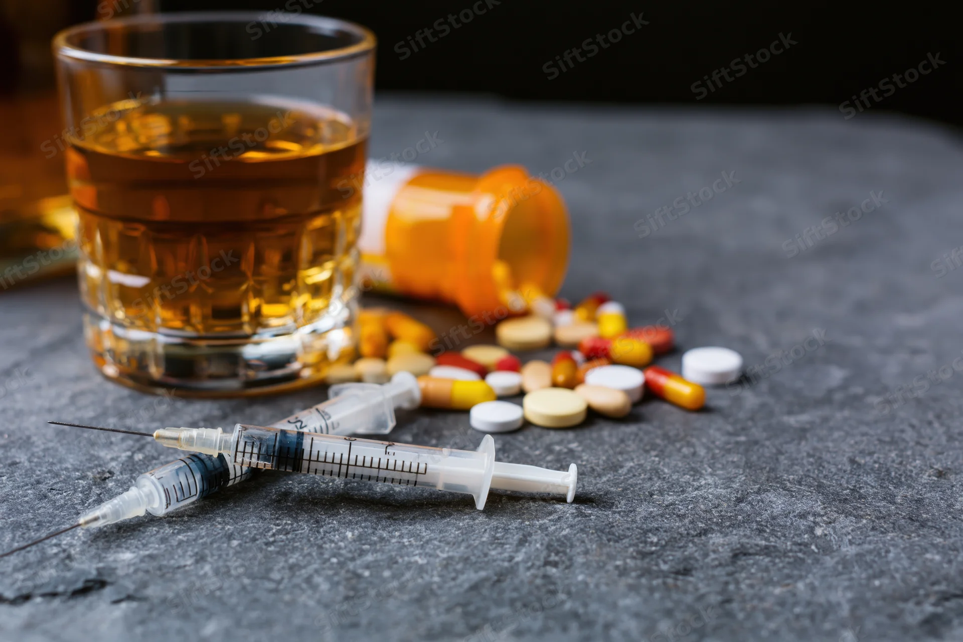 Used syringes and scattered pills beside a glass of amber liquid on a dark stone surface