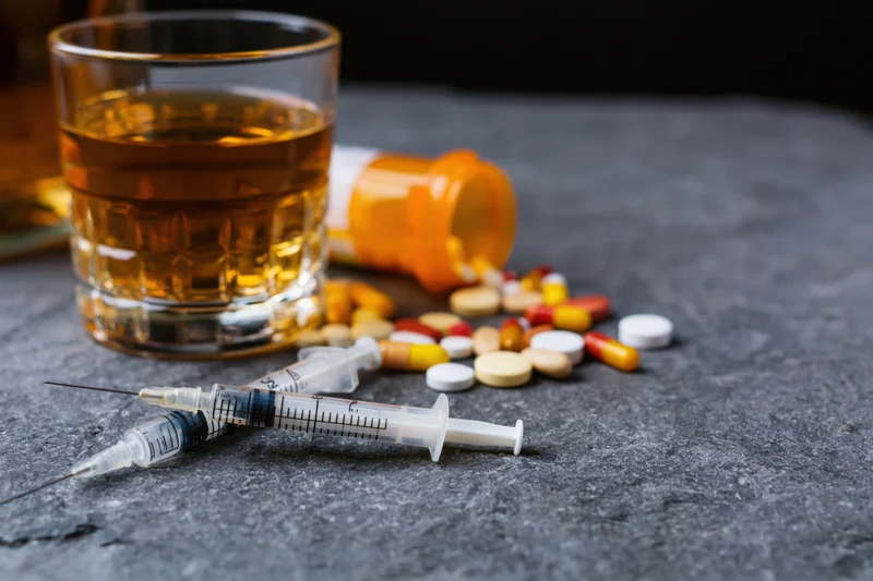 Used syringes and scattered pills beside a glass of amber liquid on a dark stone surface