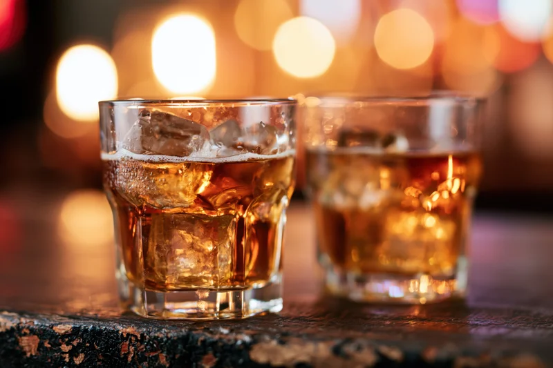 Two whiskey glasses filled with amber liquid and ice on a wooden bar surface at night