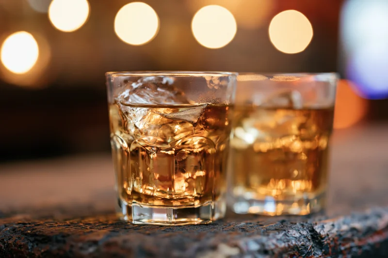 Two whiskey glasses with ice on a stone surface in a dimly lit bar setting