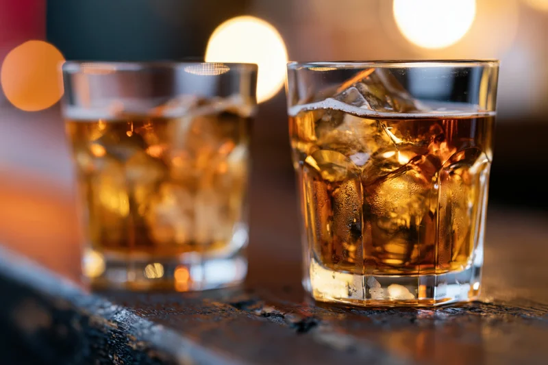 Two whiskey glasses with ice on a dark wooden bar surface under warm ambient lighting