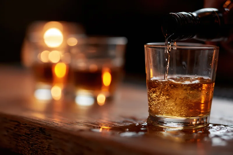 Whiskey being poured from a dark bottle into a rocks glass on a wooden table with warm candlelight in the background