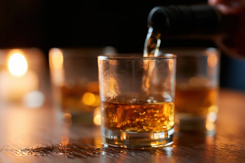 Whiskey being poured into a rocks glass on a wooden table with candlelight in the background
