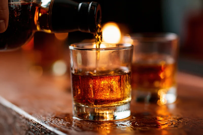 Whiskey being poured from a dark bottle into a rocks glass on a wooden bar with condensation and warm lighting