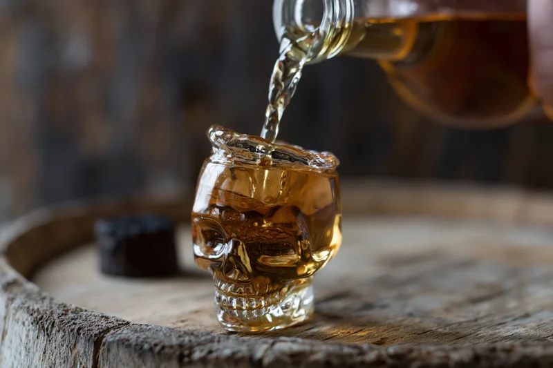 Whiskey being poured from a bottle into a skull-shaped glass on a wooden barrel top