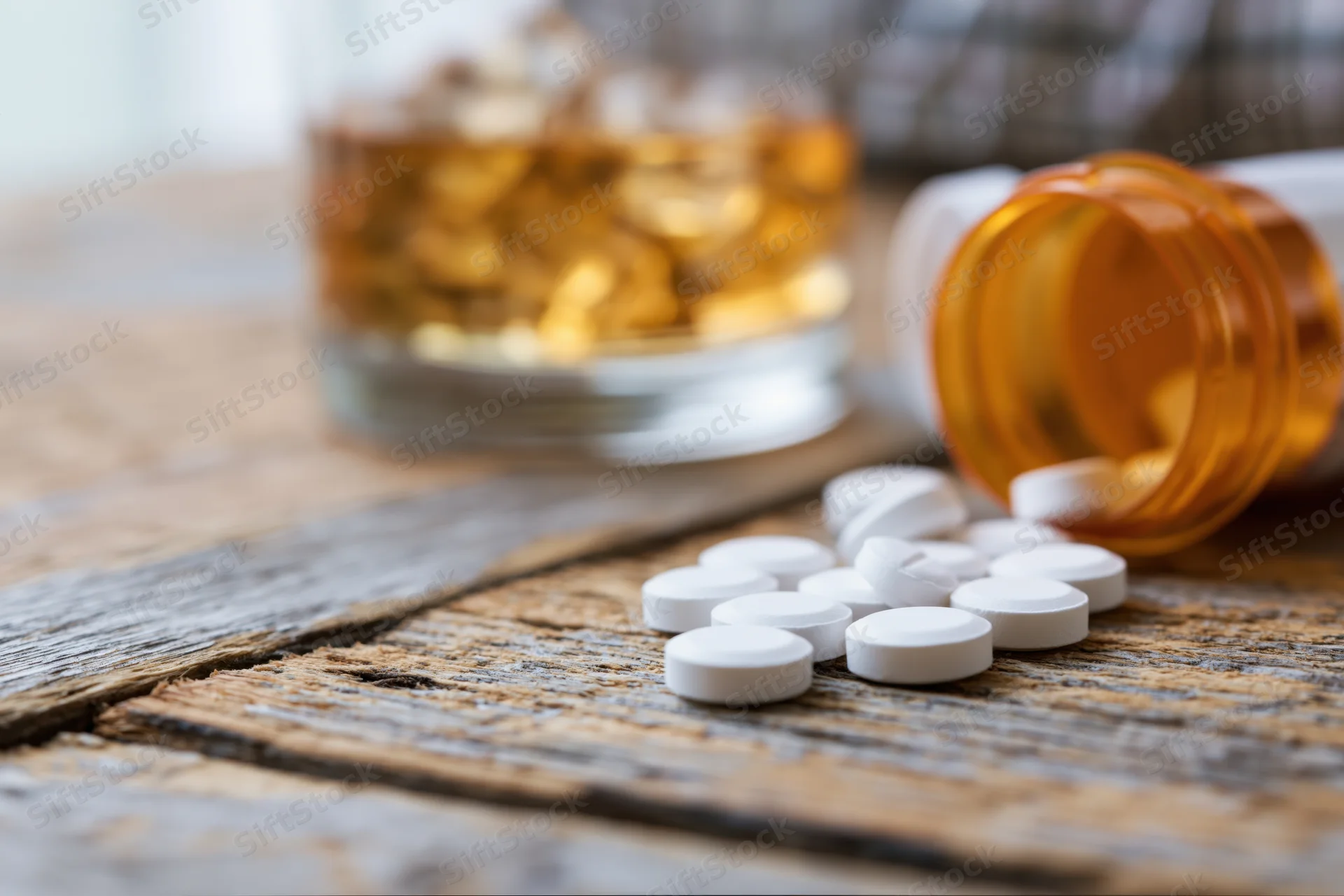 White pills spilled from an orange bottle on a worn wooden table beside a glass of amber liquid