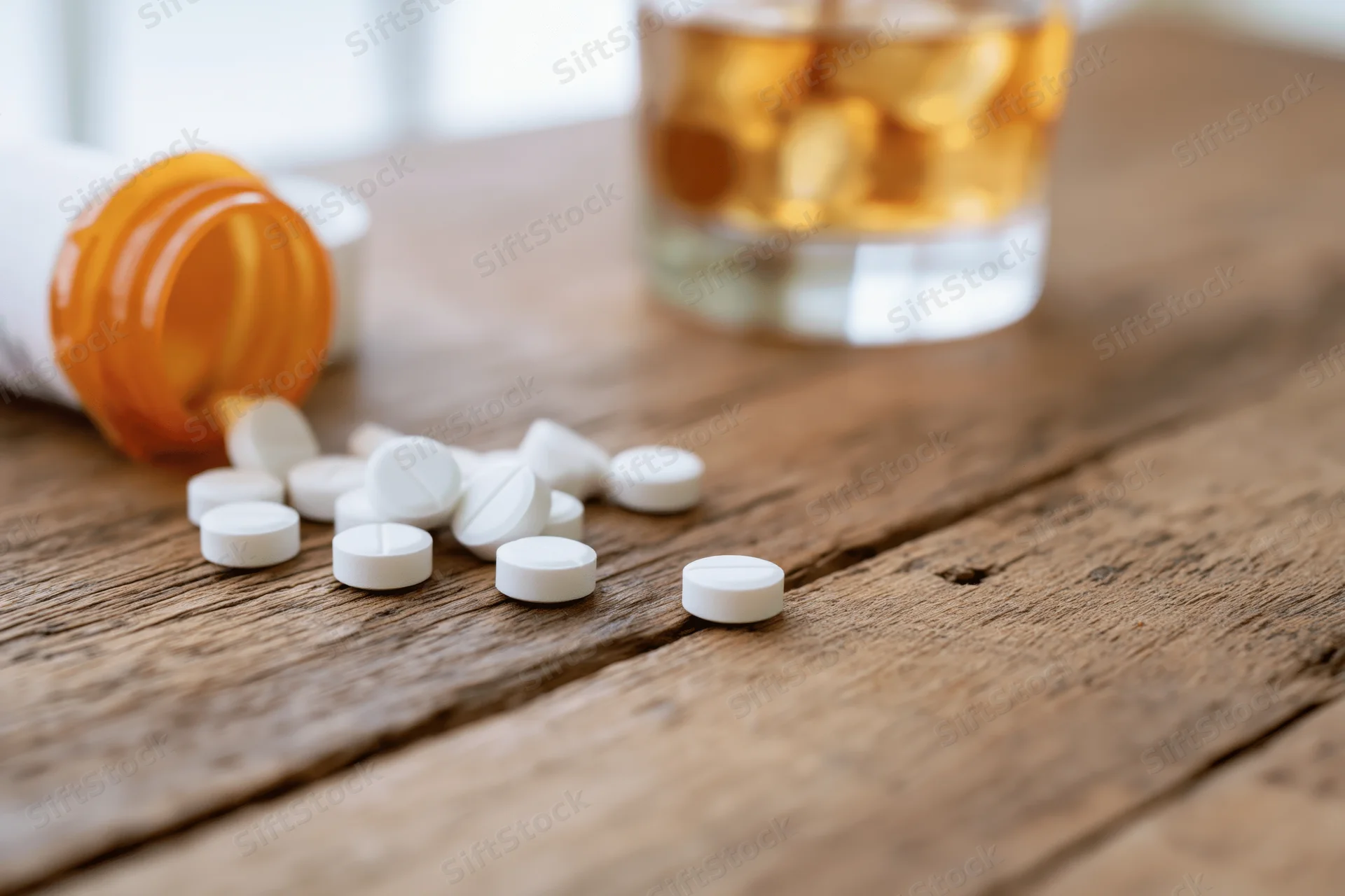 White pills scattered on a wooden table beside a glass of whiskey and a tipped pill bottle