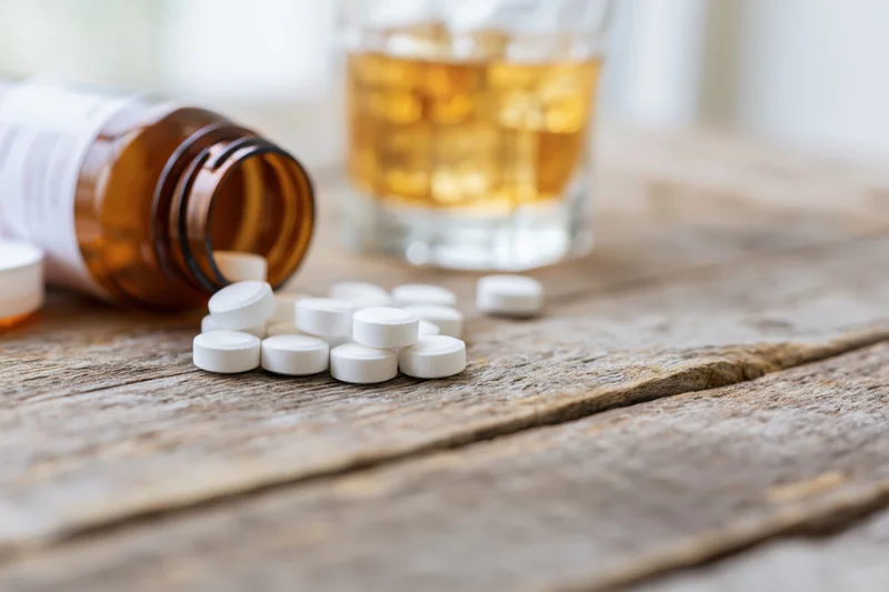 White pills scattered on a worn wooden table beside a glass of whiskey