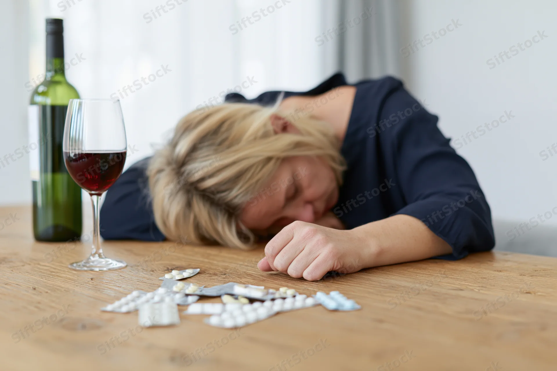 Woman with blonde hair slumped forward with head resting on wooden table beside wine glass and scattered pills