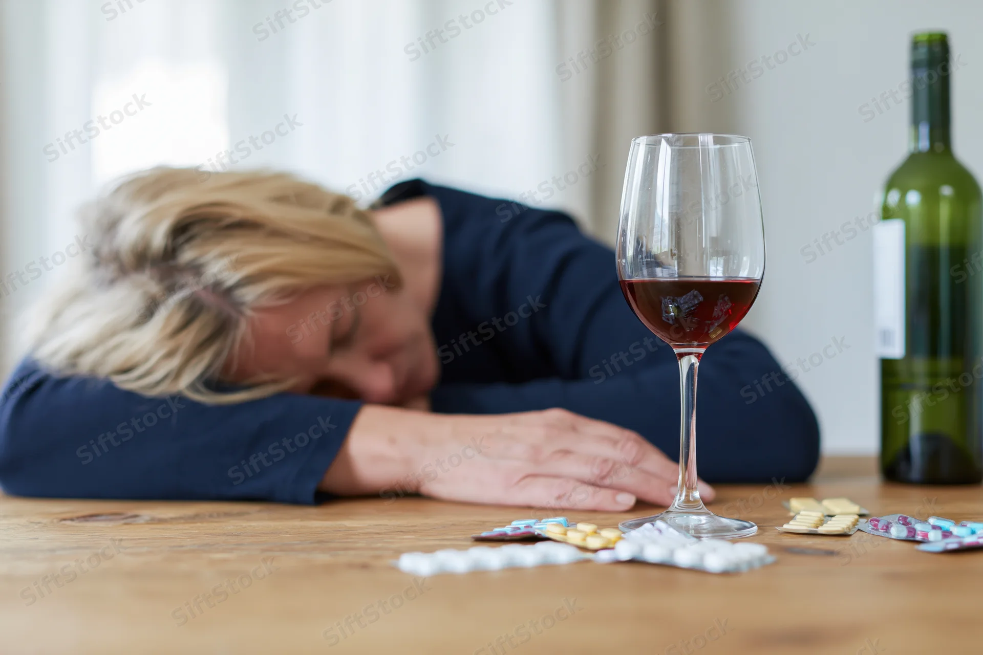 Woman with blonde hair slumped forward resting head on wooden table beside wine glass and scattered pills