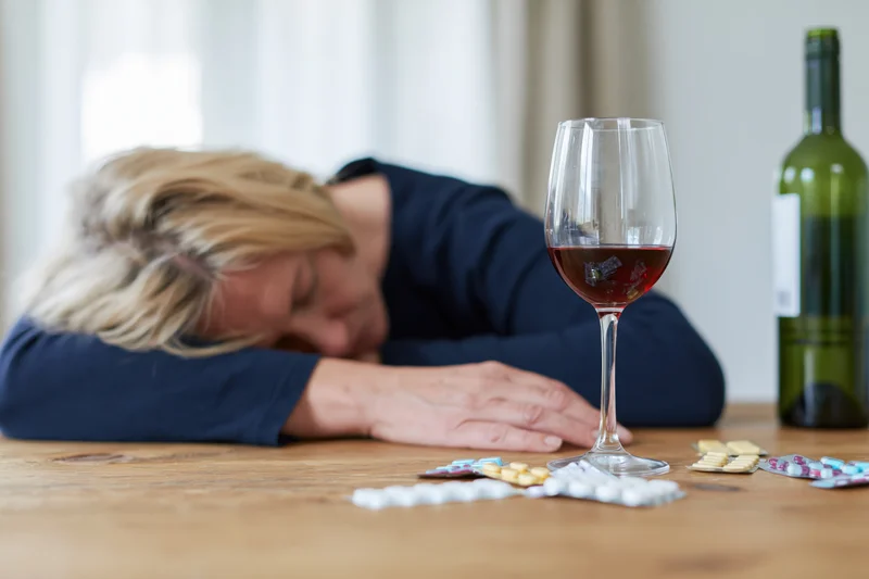 Woman with blonde hair slumped forward resting head on wooden table beside wine glass and scattered pills