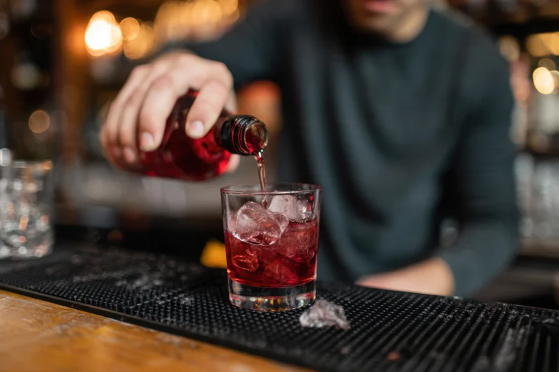 Bartender pouring red liquor into a glass with ice on a bar counter