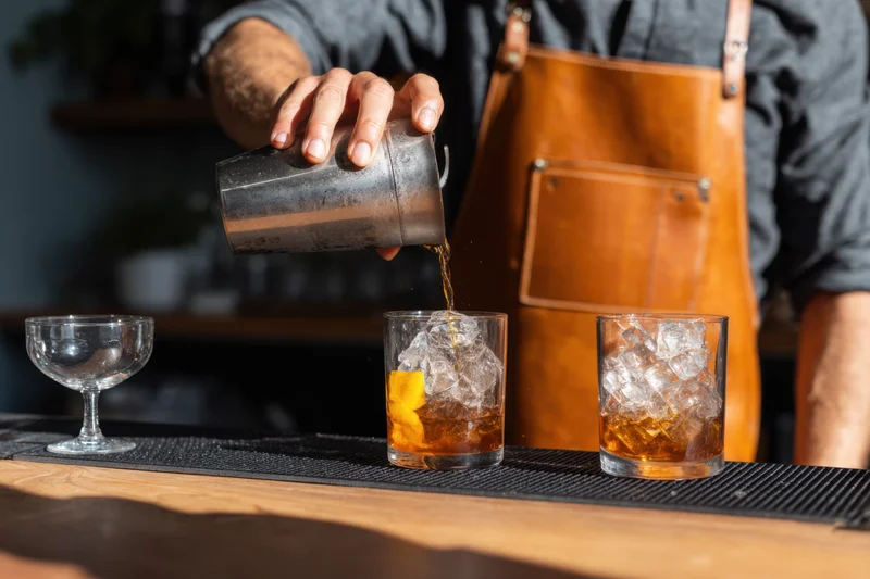 Bartender pouring whiskey into a glass with ice and orange garnish on a bar counter