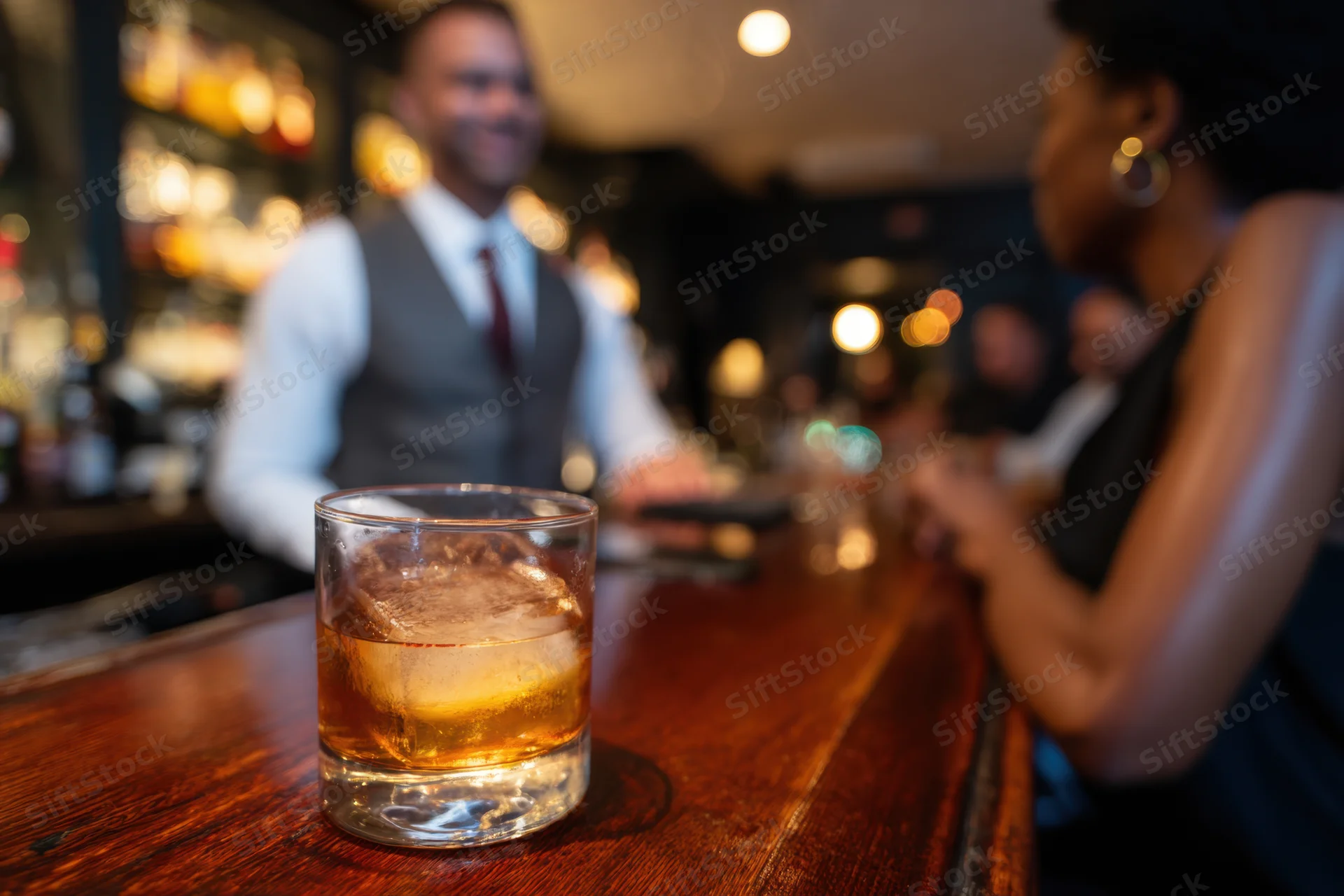 Whiskey glass with ice on wooden bar counter, bartender and woman in background, dimly lit bar interior