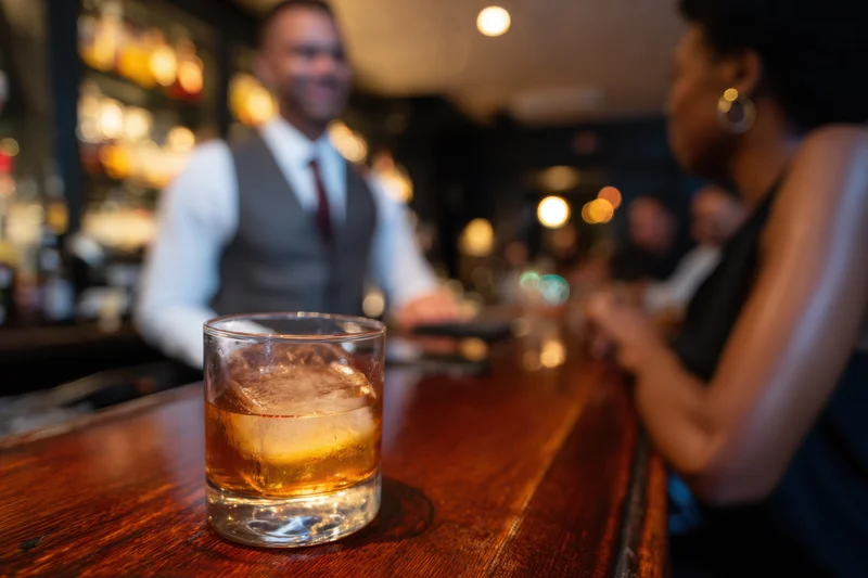 Whiskey glass with ice on wooden bar counter, bartender and woman in background, dimly lit bar interior