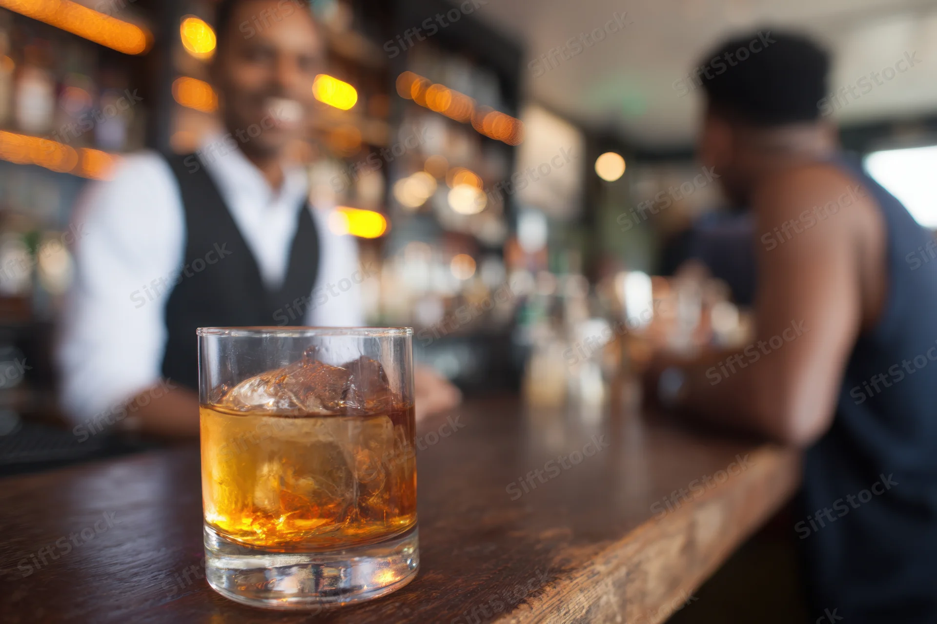Whiskey glass with ice on wooden bar counter, bartender and customer in background
