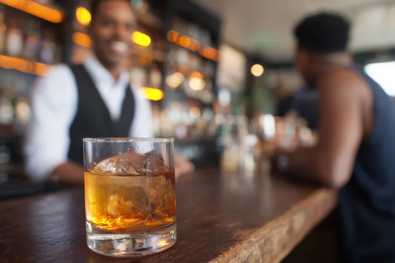 Whiskey glass with ice on wooden bar counter, bartender and customer in background