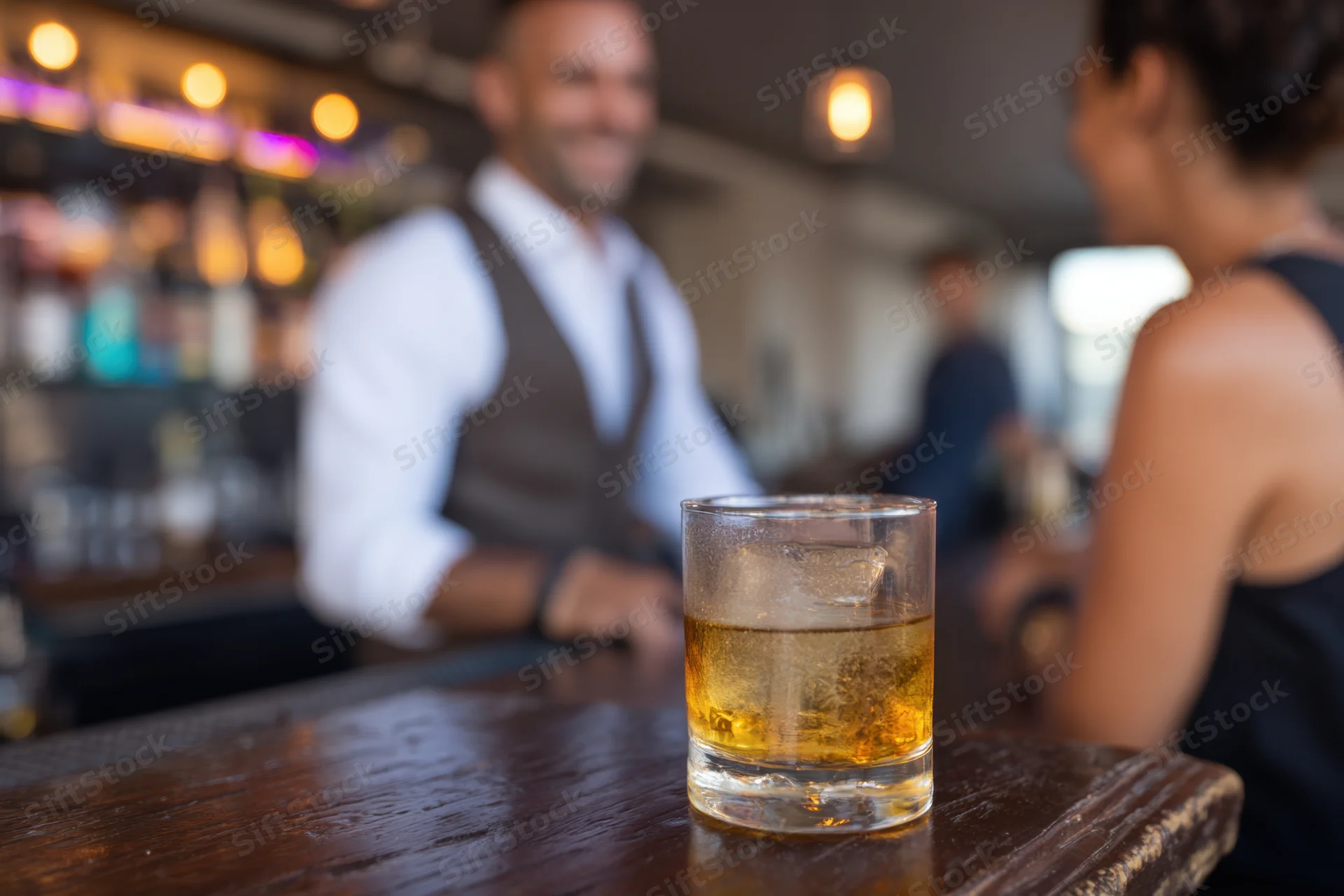 Whiskey glass with ice on wooden bar counter, bartender and woman in background conversing